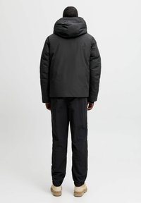 Sort pufferjakke med hætte, har lynlås lukning og let cropped design. Bæres med sorte, løstsiddende bukser og beige sko.