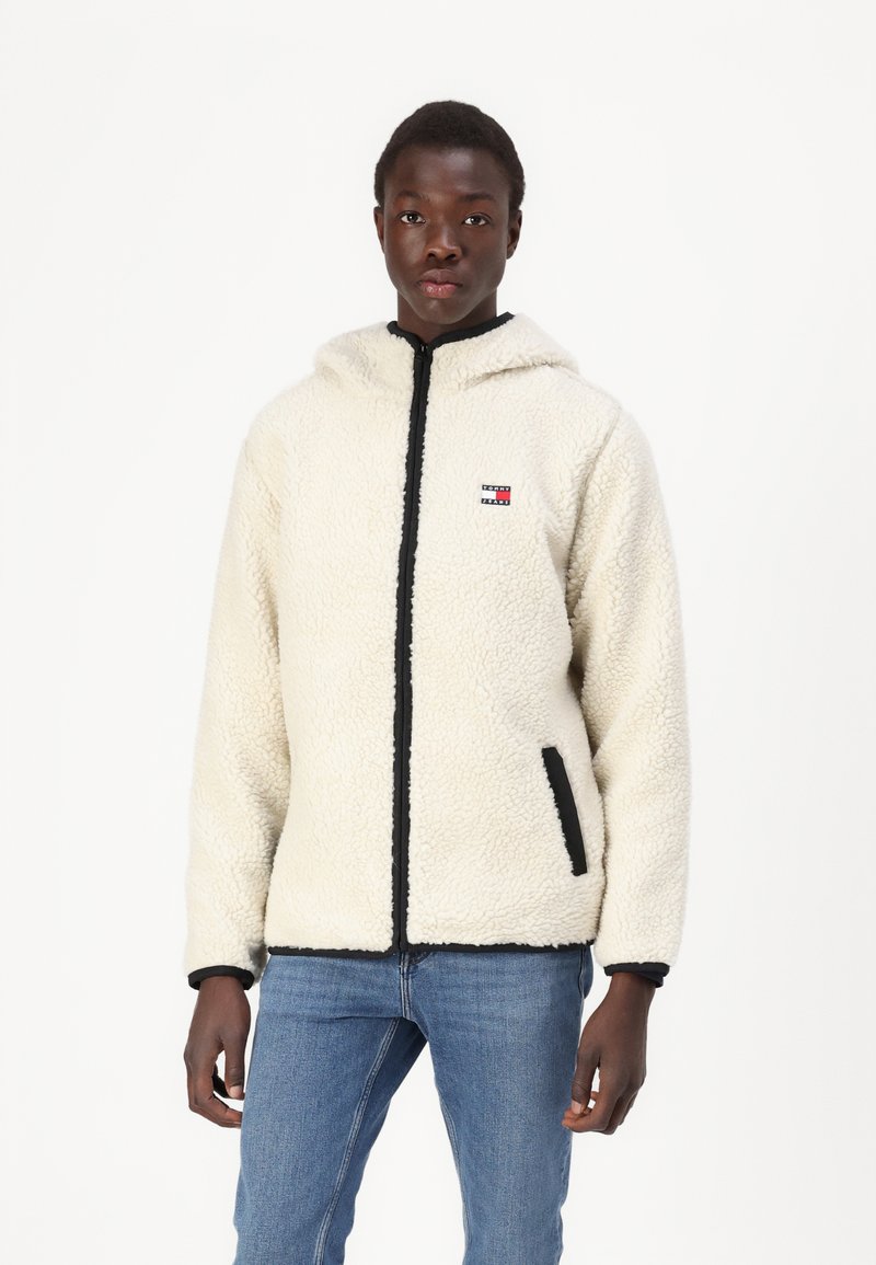 Veste en polaire synthétique crème avec des bordures noires, fermeture éclair, poches latérales et un patch logo sur la poitrine. Dispose d'une capuche ajustée.