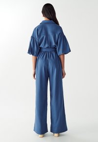 Combinaison bleue avec un col, manches courtes bouffantes, taille cintrée et pantalons évasés, fabriquée à partir d'un tissu doux.