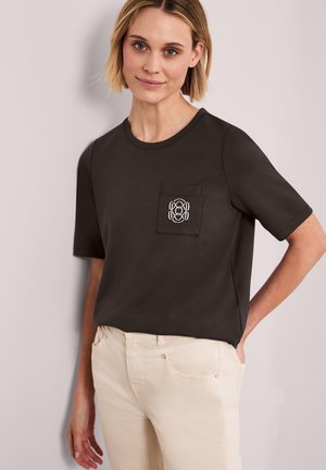 Frau trägt ein dunkelbraunes Kurzarmshirt mit Tasche und gesticktem Logo, kombiniert mit hellbeigen Hosen und steht vor einer schlichten Wand.
