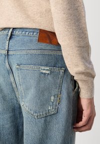 Lys blå denimbukser har en falmet tekstur, slitte detaljer på baklommen og et brunt lærdekor på livet.