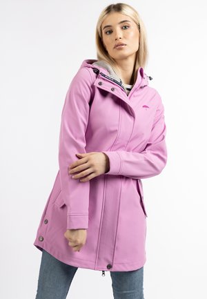Donna con capelli biondi che indossa un impermeabile rosa chiaro con cappuccio e jeans blu, in piedi davanti a uno sfondo bianco neutro.