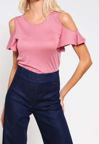 Pink cold-shoulder top with ruffle sleeves, made of soft fabric, paired with high-waisted navy trousers featuring a front button closure.