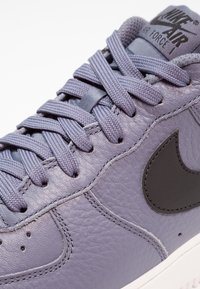 Buty Nike Air Force 1 w matowej lawendowej skórze z czarnymi akcentami, z płaskimi sznurowadłami, wyściełanym językiem oraz teksturowanymi detalami na powierzchni.