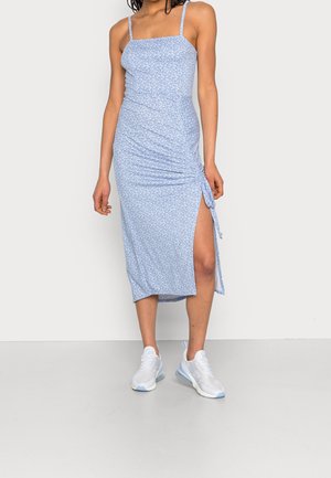 Femme portant une robe midi fleurie bleu clair avec fines bretelles et fente latérale, associée à des baskets blanches de sport.