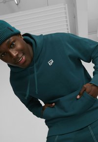 Sudadera de algodón verde azulado con un bolsillo frontal y capucha con cordón, presenta un pequeño logotipo blanco de Puma. Detalles de puños y dobladillo acanalados.