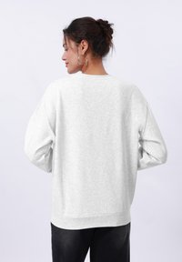 Felpa grigia chiara con colletto a girocollo, spalle scese e polsini a costine. Tessuto morbido, vestibilità rilassata e design minimalista.