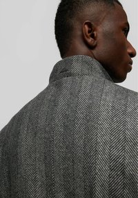 Veste grise à motif chevron présentant un col montant, avec un tissu texturé et un design structuré. Détail notable à l'arrière près du col.