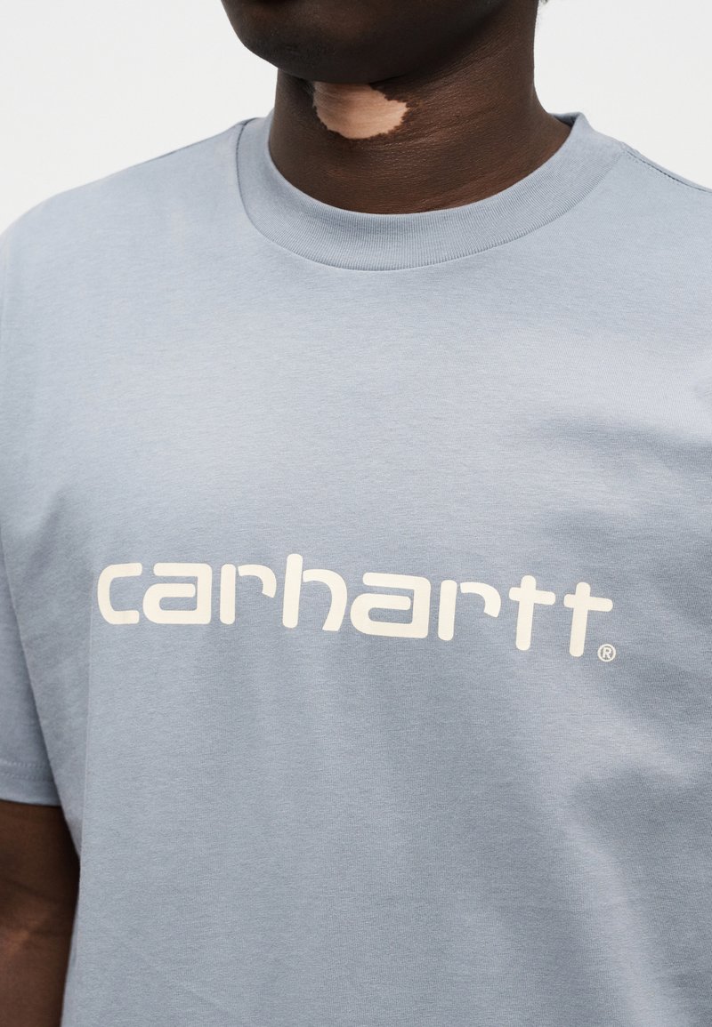 Lichtblauw katoenen T-shirt met een groot wit "carhartt" logo in een modern lettertype, korte mouwen en een ronde hals.