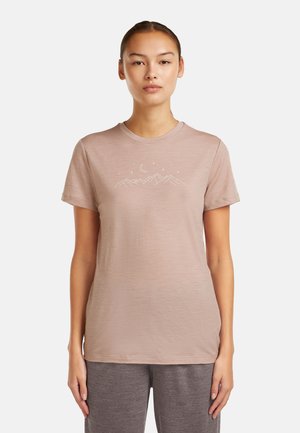 Woman wearing a light mauve t-shirt with a white mountain, moon, and stars design, paired with gray pants, standing against a plain background.