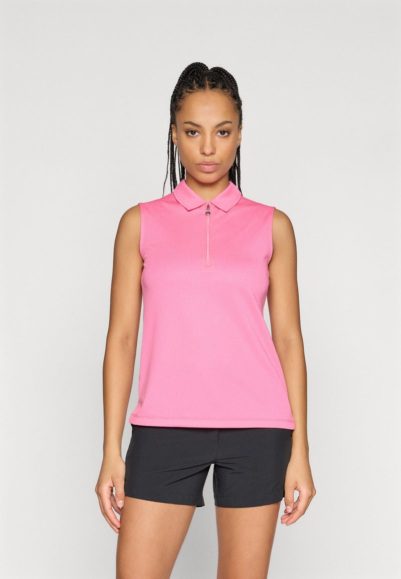 Polo-skjorte i klar rosa, korte ermer, glidelås foran, teksturert stoff. Kombinert med svarte shorts, modell står mot en ensfarget bakgrunn.