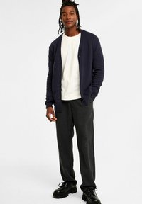 Dunkelblaues Cardigan mit gerippten Ärmeln und Knopfverschluss, getragen über einem weißen T-Shirt und kombiniert mit grauen Hosen und schwarzen Schnürschuhen.