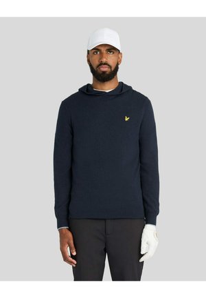 Homme portant un sweat à capuche bleu marine avec un logo d'oiseau jaune, pantalon noir, gant de golf blanc à la main droite et casquette blanche perforée, debout devant un fond gris.