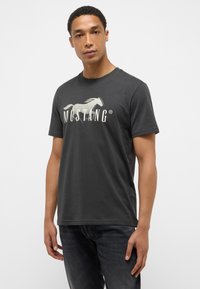 Camiseta de algodón gris oscuro con un gran logo de caballo impreso y el texto "MUSTANG" en color crema. Manga corta y cuello redondo.
