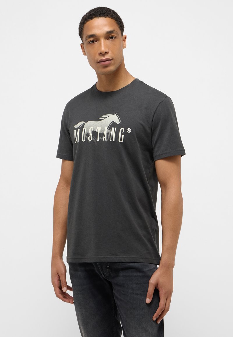 Camiseta de algodón gris oscuro con un gran logo de caballo impreso y el texto "MUSTANG" en color crema. Manga corta y cuello redondo.