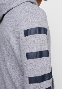 Grå sweatshirt i en texturerad väv. Har marinblå glansiga ränder på ärmarna som skapar en kontrast mot basfärgen.