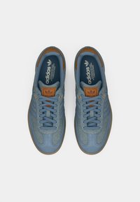 Ζευγάρι μπλε αθλητικά παπούτσια Adidas με καφέ δερμάτινες λεπτομέρειες και μπλε κορδόνια, σε προβολή από πάνω σε λευκό φόντο.