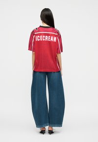 Γυναίκα με μακριά σκούρα μαλλιά, φορώντας κόκκινη κοντομάνικη φανέλα με την επιγραφή "ICECREAM" και ευρύχωλο μπλε τζιν παντελόνι, στέκεται με την πλάτη σε απλό φόντο.