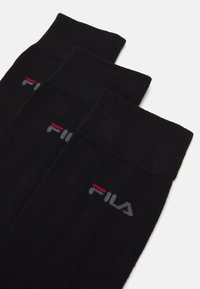 Trois paires de chaussettes noires à mi-mollet avec des logos FILA gris et rouge sur le revers, disposées en chevauchement sur un fond blanc.