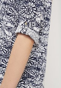 Navy and white patterned blouse with floral designs. Features rolled-up sleeves secured with gold button. Lightweight fabric with a smooth texture.