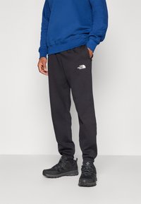 Sorte sweatpants med elastiske manchetter, med et hvidt logo på venstre lår. Parret med en blå sweatshirt og sorte sportsko.