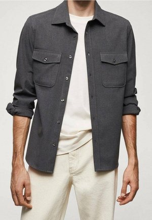 Mężczyzna w ciemnoszarej koszuli typu overshirt z zapinanymi kieszeniami, białym t-shirtem i kremowych dżinsach. Podwinięte rękawy, pokazany od ramion do połowy uda.