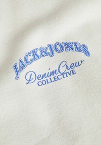 Tessuto beige con testo ricamato blu che recita "JACK & JONES Denim Crew COLLECTIVE", che mette in mostra una superficie liscia.