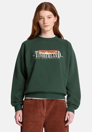 Jonge vrouw met lang rood haar draagt een donkergroene Timberland trui en een bruine ribfluwelen broek, staand tegen een effen achtergrond.