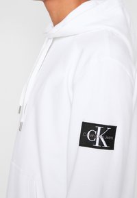 Vit bomulls-Hoodie med justerbar dragsko, med en lapp med "CK"-logotyp och texten "Calvin Klein Jeans" på vänster ärm.