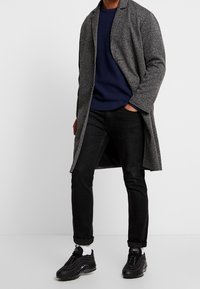 Manteau gris léger à finition texturée, porté sur un pull côtelé bleu marine, associé à un jean noir et des chaussures de sport noires.