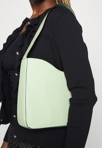 Femme portant un cardigan noir à volants et tenant un sac à bandoulière vert clair sur son épaule droite, sur un fond uni.