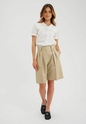 Shorts plissés beiges à taille haute, dotés de deux boutons, associés à une chemise blanche à manches courtes cintrée. Des chaussures noires complètent le look.
