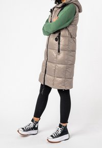 Osoba ubrana w bezrękawnik z długim szarym wypełnieniem, zieloną koszulę z długim rękawem, czarne legginsy z prążkowanej tkaniny oraz czarne sneakersy na platformie z wysoką cholewką.