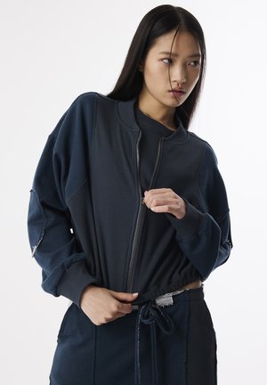 Giacca zip-up navy con texture a costine, vestibilità oversize e orlo cropped. Presenta una zip liscia e dettagli sdruciti sulle maniche.