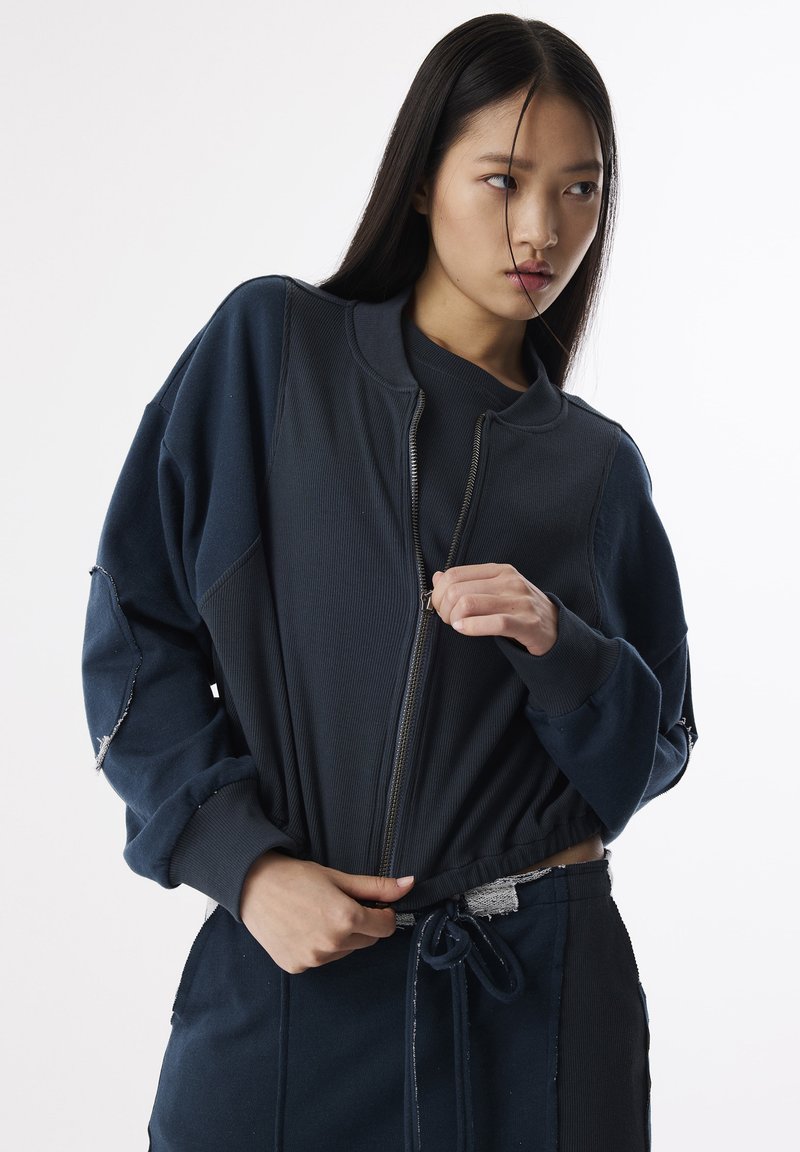 Marine blauwe zip-up jack met geribbeld textuur, oversized pasvorm en een korte zoom. Voorzien van een soepele rits en versleten accenten op de mouwen.