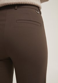 Bruna skräddarsydda byxor i ett slätt tyg, med en bakficka och en cirkulär metalldetalj vid midjan.