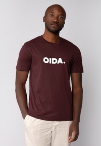 Bordeauxfarbene Baumwoll-T-Shirt mit kurzen Ärmeln und rundem Halsausschnitt. Mit großen weißen Buchstaben "OIDA." auf der Brust. Schlichtes Design mit minimalen Details.
