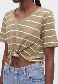 Femme portant un t-shirt court beige et blanc à rayures en V noué devant avec un jean taille haute bleu clair.