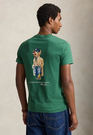 Personne portant un t-shirt vert avec un graphisme au dos représentant un ours en manteau et casquette, associé à un jean bleu, debout de profil sur un fond uni.