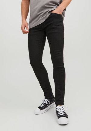 Hombre que lleva jeans negros ajustados, zapatillas en blanco y negro, y una camisa gris, de pie con una mano en el bolsillo frente a un fondo liso.