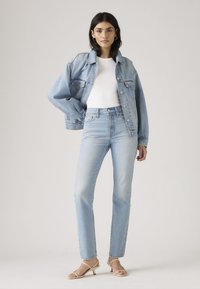 Lichtblauwe denimjas over een witte nauwsluitende top, gecombineerd met lichtblauwe spijkerbroek. Model draagt lichte sandalen. Gladde texturen, casual pasvorm.