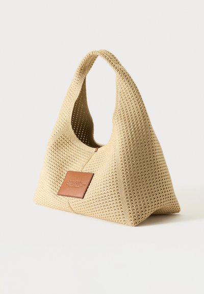 Sac à main en tissu tissé beige avec une large anse et un patch en cuir marron à l’avant, sur un fond uni clair.