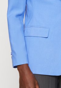 Helles blaues Blazer mit strukturiertem Design, einer einzigen Brusttasche und drei Knöpfen am Ärmel. Der Stoff wirkt glatt mit minimaler Textur.