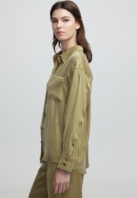Camisa de manga larga en verde oliva, confeccionada en una tela suave, con un frente abotonado, un bolsillo en el pecho y mangas con puños que tienen cierres de botones.
