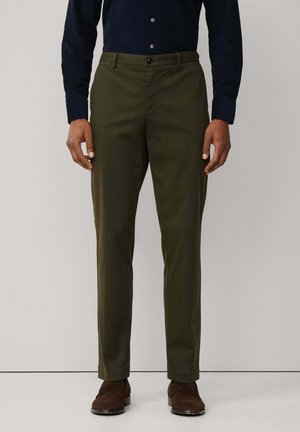Olivgrüne Hose aus glattem Stoff, mit Knopfverschluss und seitlichen Taschen. Getragen mit einem dunkelblauen Hemd mit Knöpfen und braunen Schuhen.