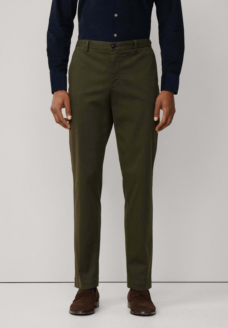 Olijfgroene broeken van gladde stof, met een knoopsluiting en zijzakken. G gedragen met een donkerblauw overhemd met knopen en bruine schoenen.