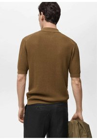 Camisa polo corta de manga con textura marrón y cuello, combinada con pantalones negros. El portador sostiene una chaqueta marrón a juego en una mano.