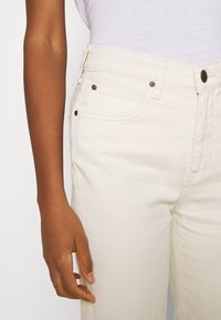 Beige denimjeans med framfickor, hög midja och metalldetaljer. Slät textur och rak bensnitt.