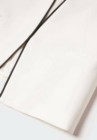 Blazer blanc avec une fine bordure noire le long du revers et des bords, avec une poche poitrine sur le côté droit.