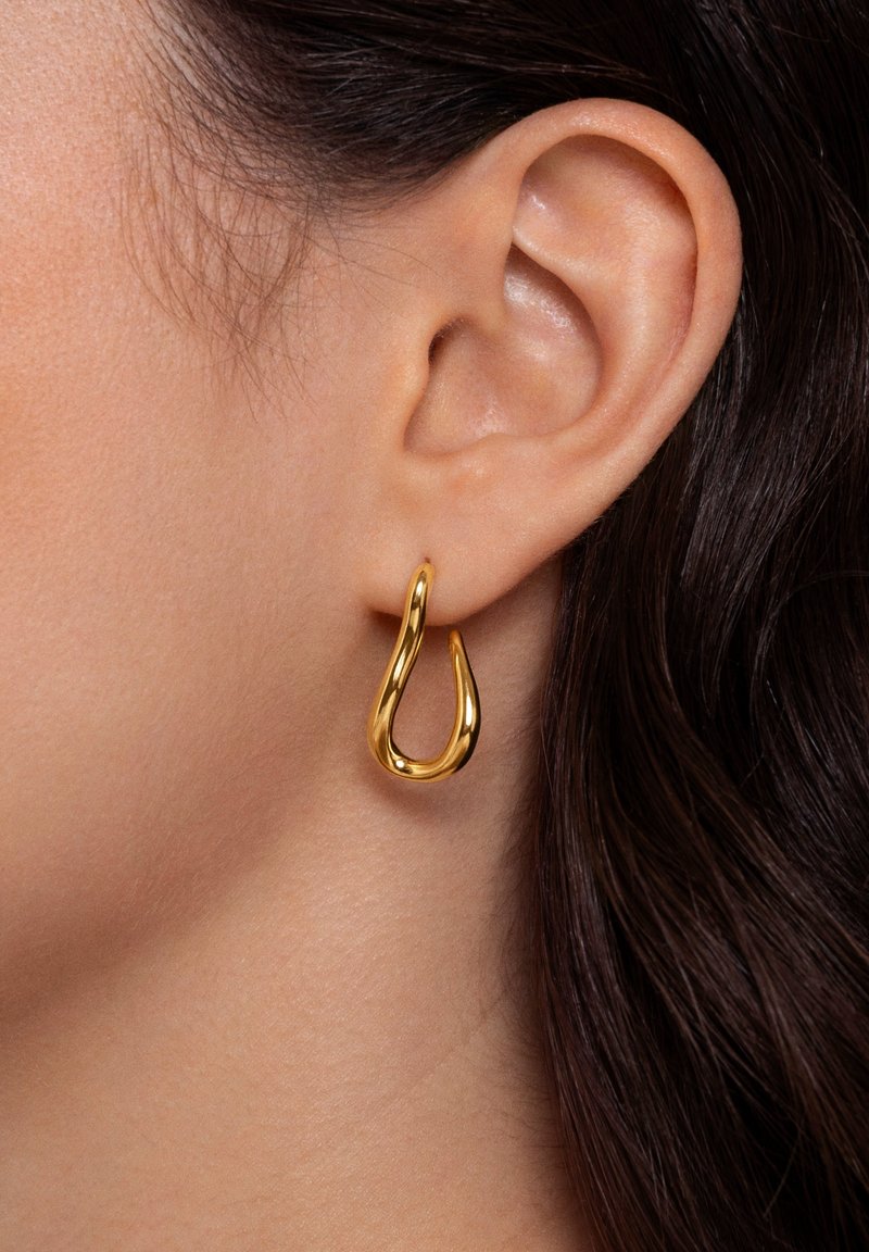 Gros plan d'une oreille gauche portant une boucle d'oreille dorée en forme de cercle brillante et irrégulière, avec des cheveux châtain foncé partiellement visibles.